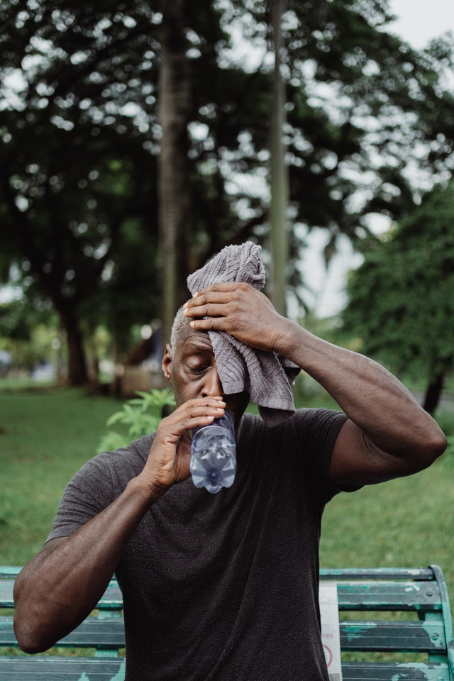 Older man in park with towel on head while drinking water.