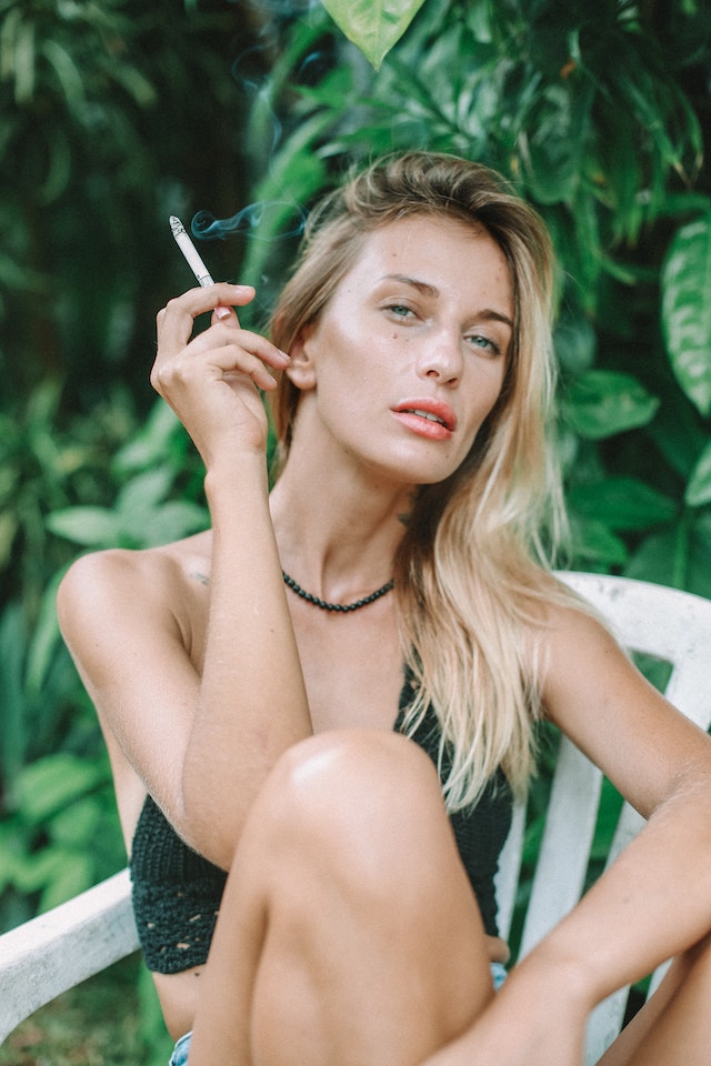 Woman sitting in lawn chair in front of greenery smoking a cigarette.