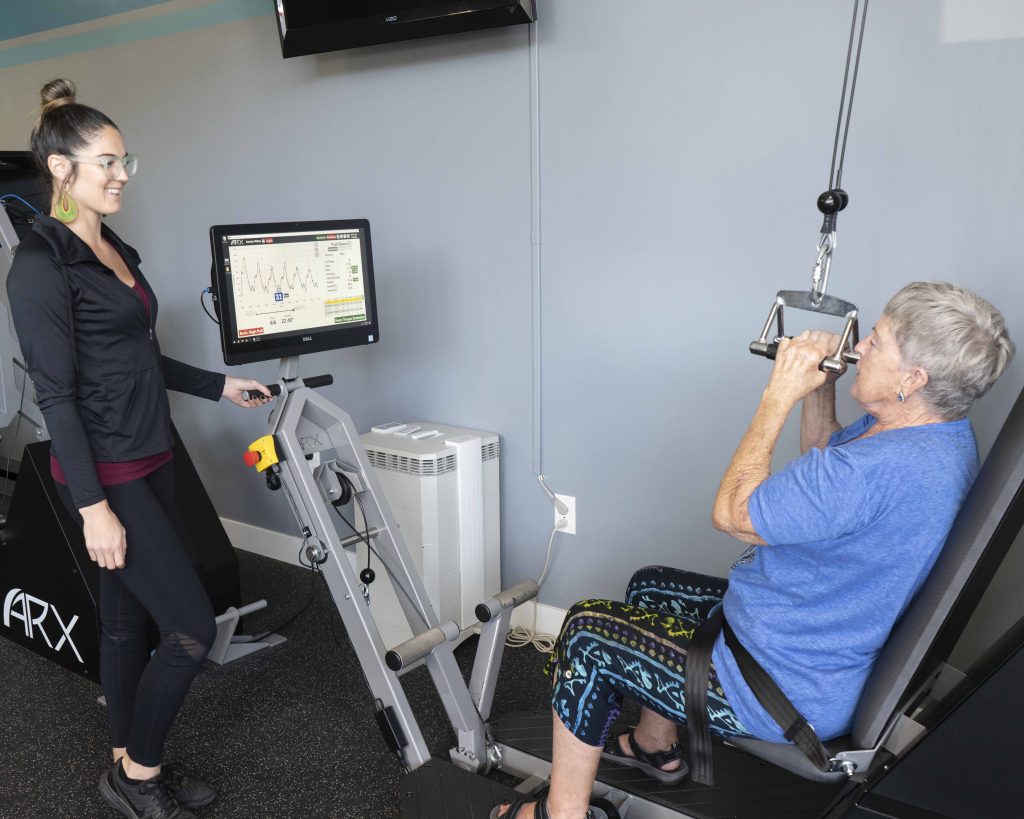 Senior woman working out on ARX machine with personal trainer at Leo's Fitness Lab.