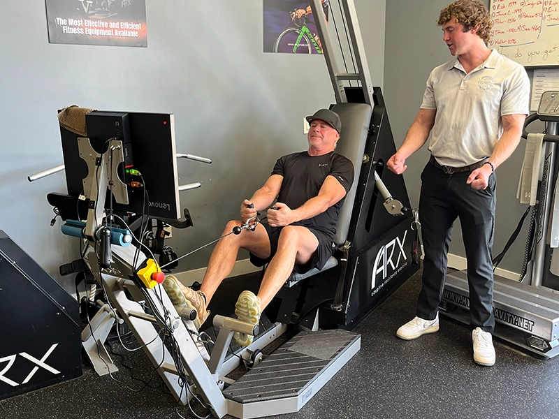 A man uses ARX fitness equipment, performing a rowing-style exercise by pulling the handles toward his torso, while a male personal trainer stands nearby, providing instruction and supervision.