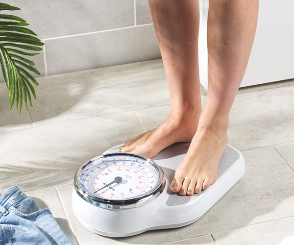 a close-up image of a person standing on a bathroom scale in the bathroom with a pair of blue jeans on the ground next to the scale