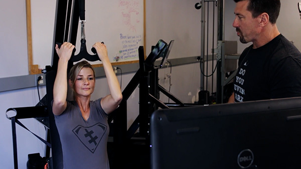 an image of a young woman using an ARX workout machine while being instructed and motivated by a personal trainer.