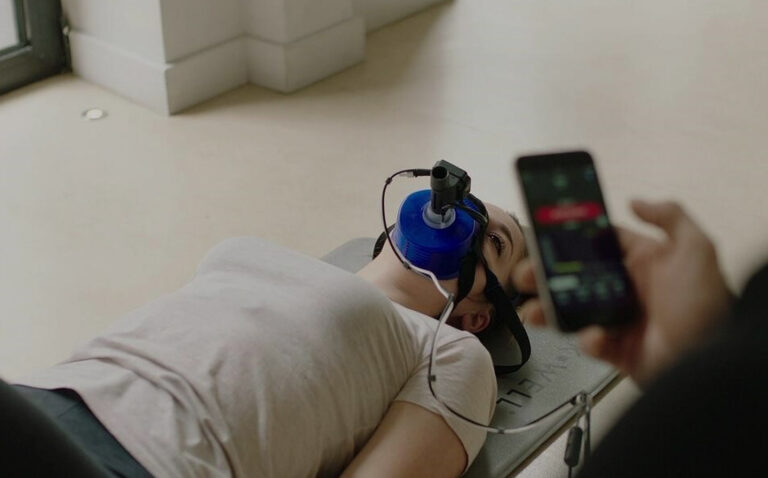 an image of a woman laying down using a precise PNOW metabolic assessment machine. a personal holding a phone is monitoring the assessment