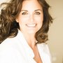 Professional headshot of a smiling woman with curly brown hair wearing a white collared shirt against a light background