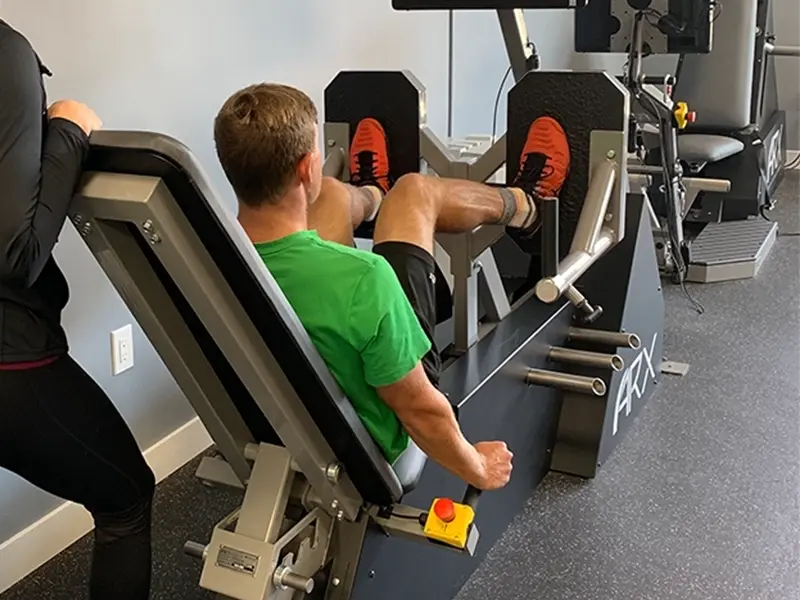 A man performs a leg press exercise on ARX fitness equipment while a female instructor stands beside him, supervising and offering guidance.